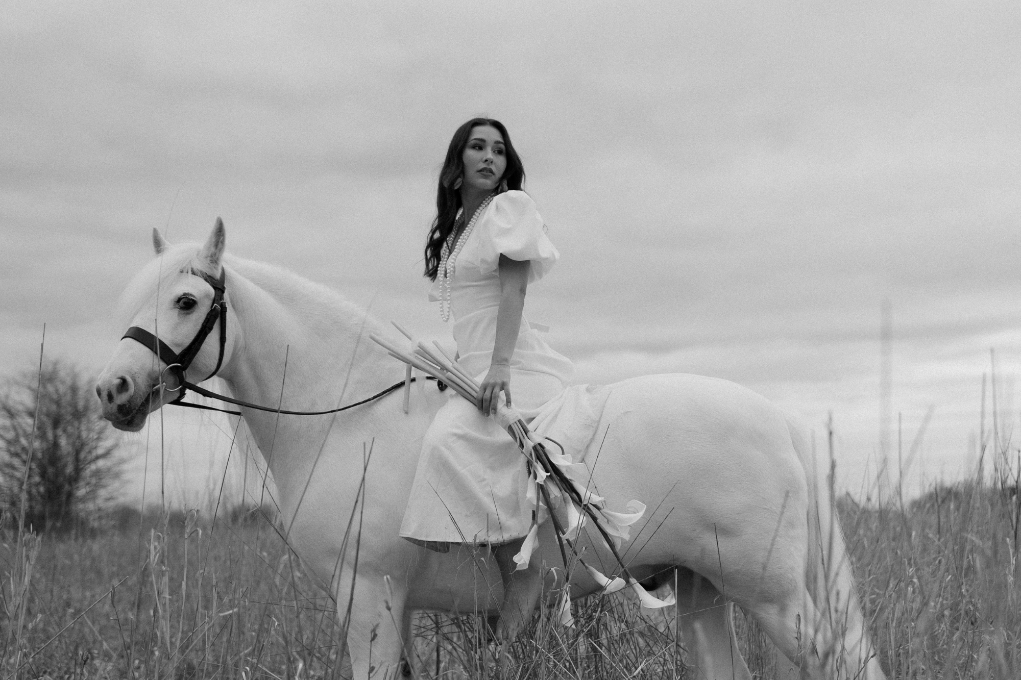 Bride holding white lilies sitting on white horse wearing pearl necklace.